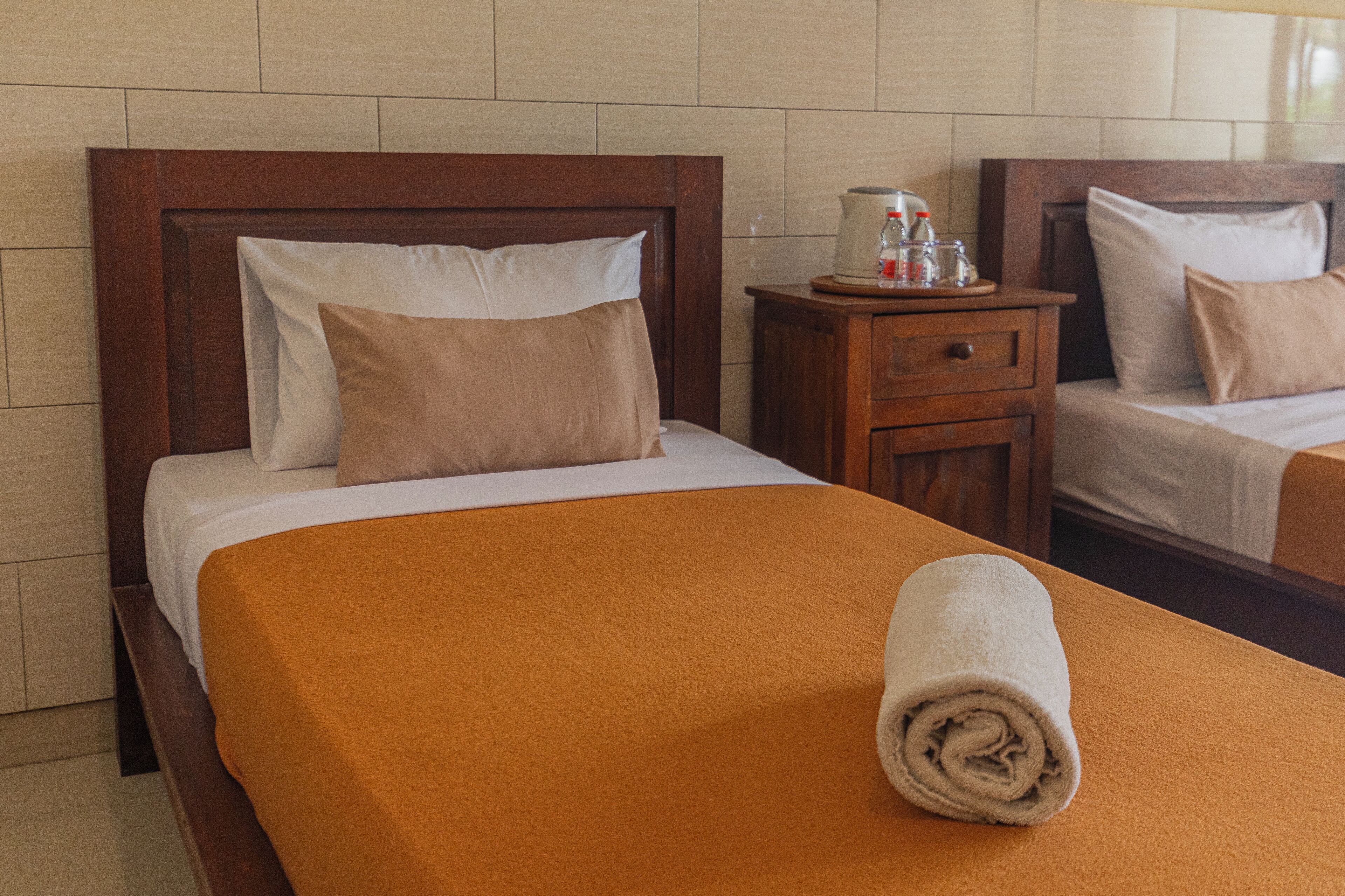 Ein Hotelzimmer mit zwei Betten, einem hölzernen Kopfteil, einem Nachttisch mit Kaffeemaschine und einem auf dem Bett gerollten Handtuch.