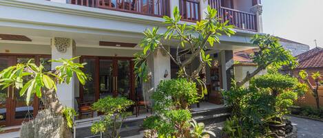 A two-story building with a balcony, surrounded by various plants and trees.