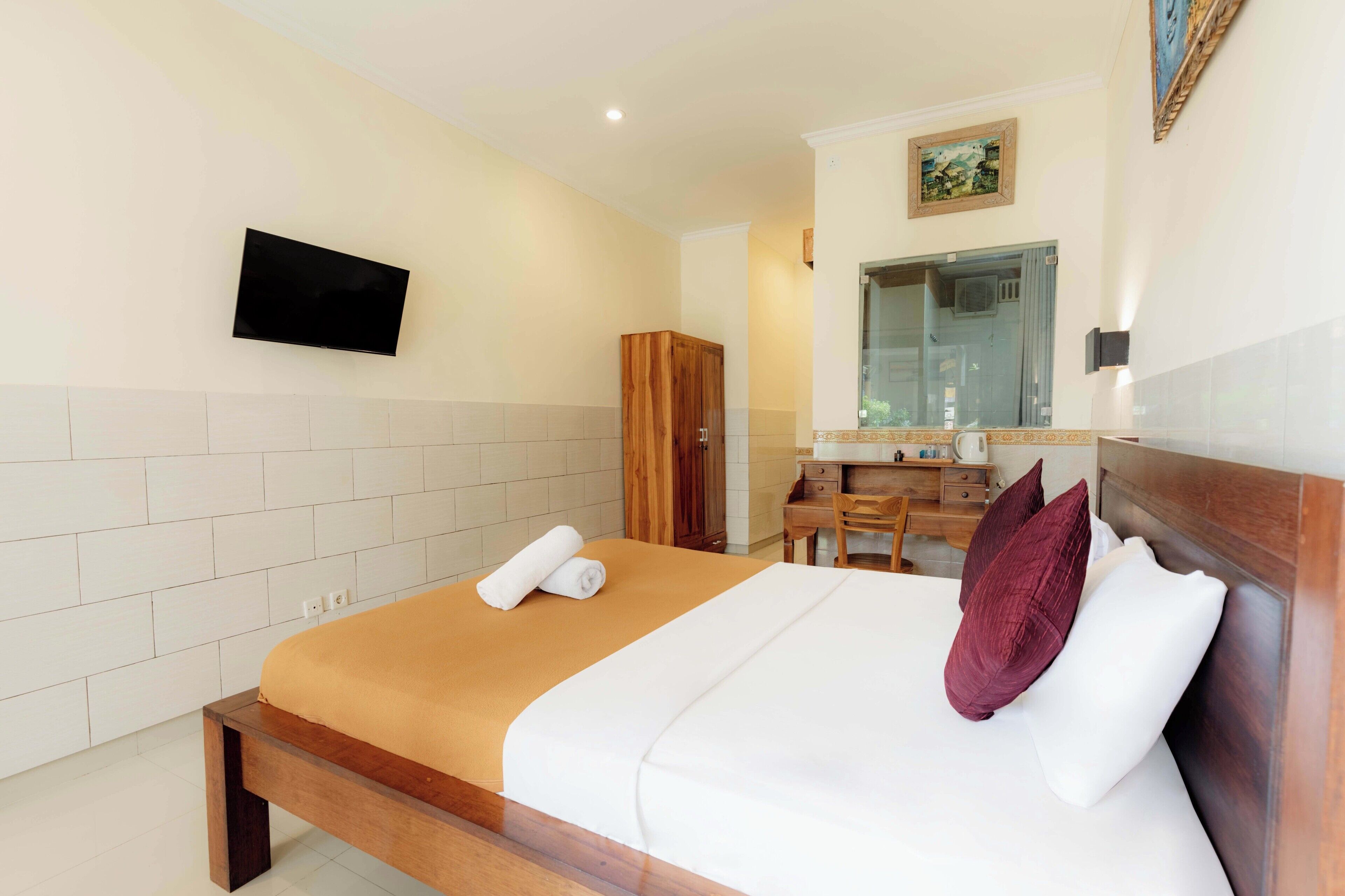 A hotel room with a bed, a wooden wardrobe, a desk with a chair, a mirror, and a television on the wall.