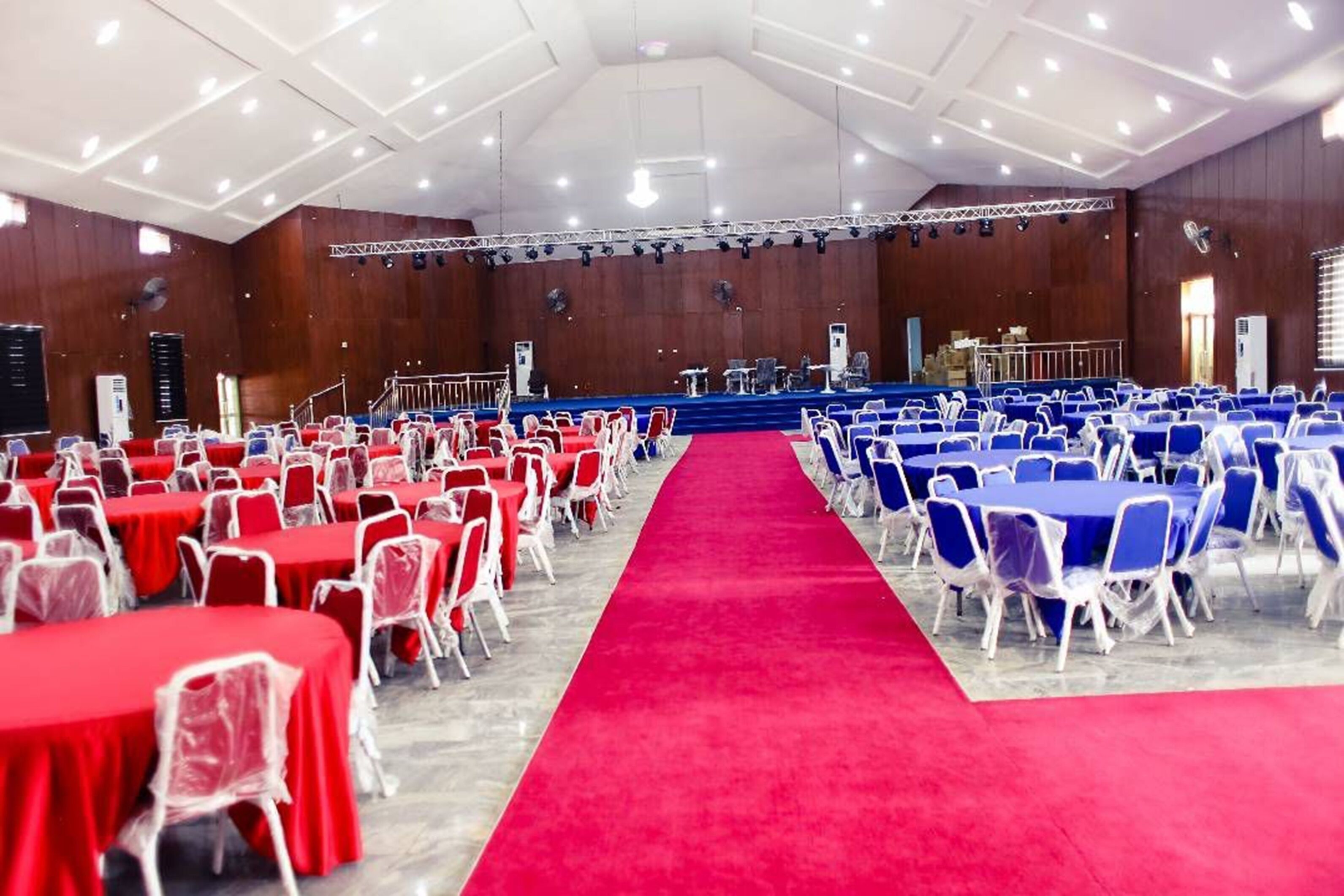 Une grande salle avec des rangées de tables et de chaises, une moquette rouge et une scène équipée d’instruments de musique.