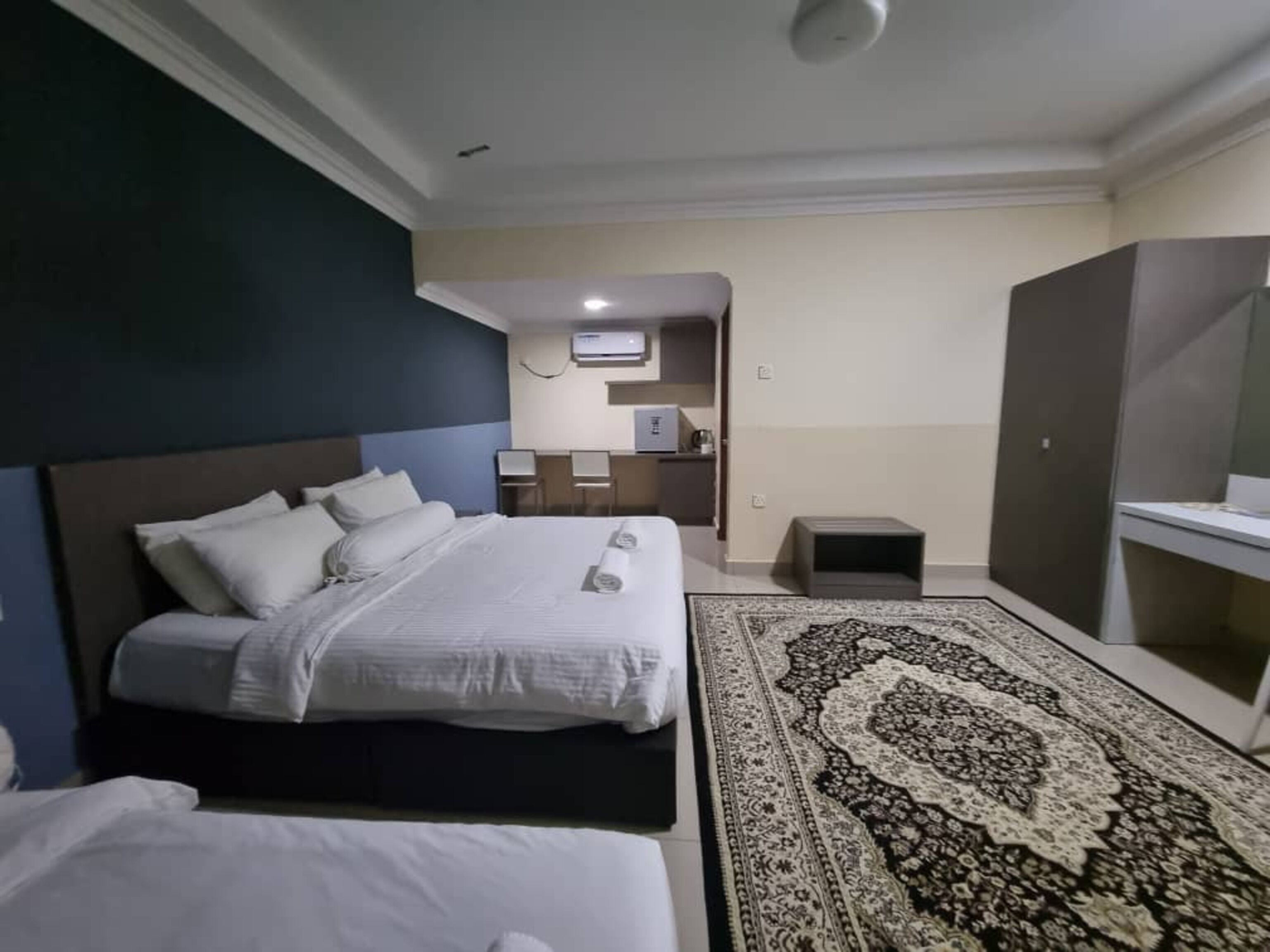 Une chambre d’hôtel avec un grand lit, un lit plus petit, un bureau avec une chaise, une armoire et un tapis à motifs.
