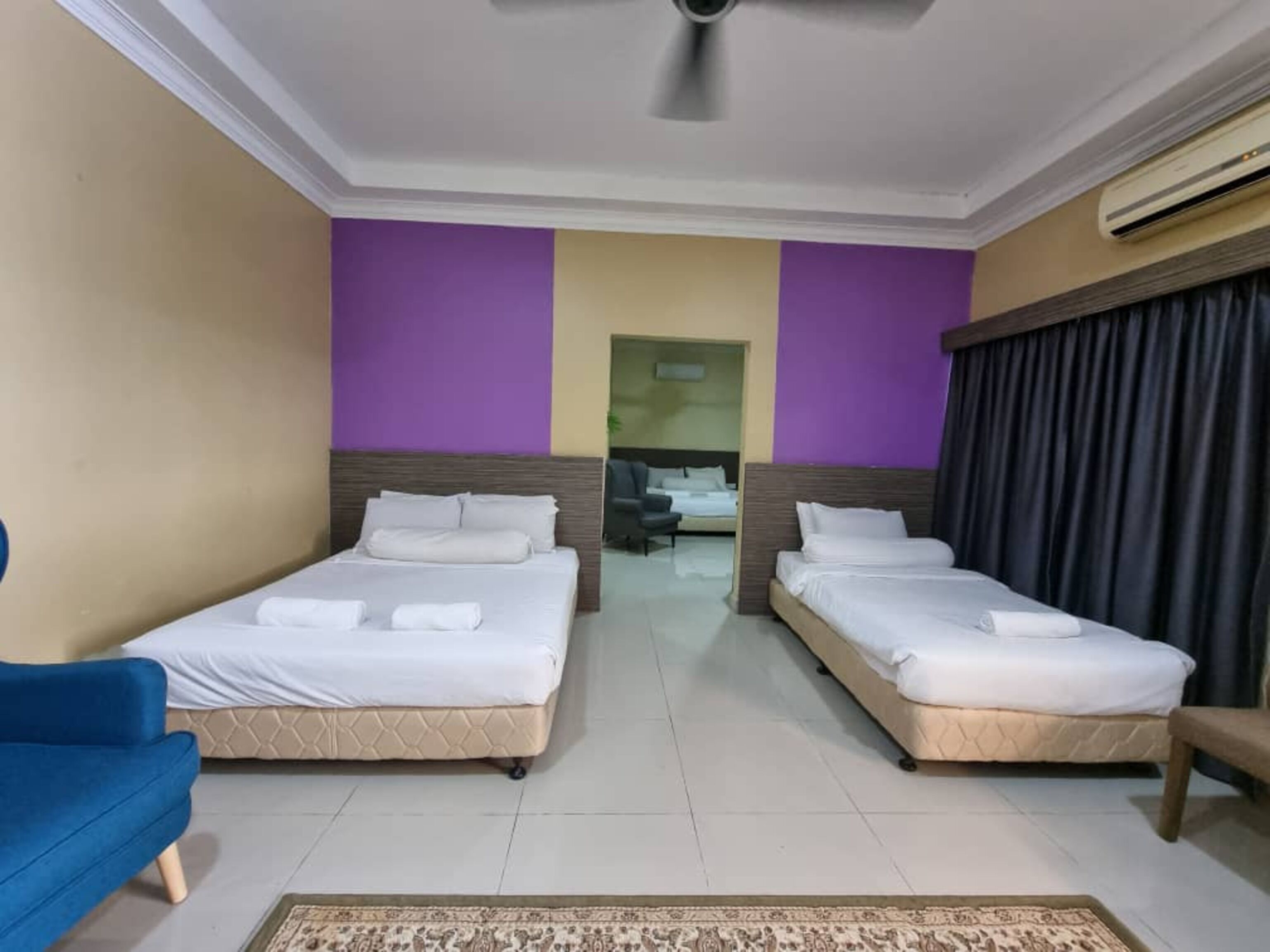A hotel room with two beds, a blue chair, a ceiling fan, and an air conditioning unit.