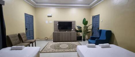 Habitación de hotel con dos camas individuales, televisor, ventilador de techo, una planta grande y un sillón azul.