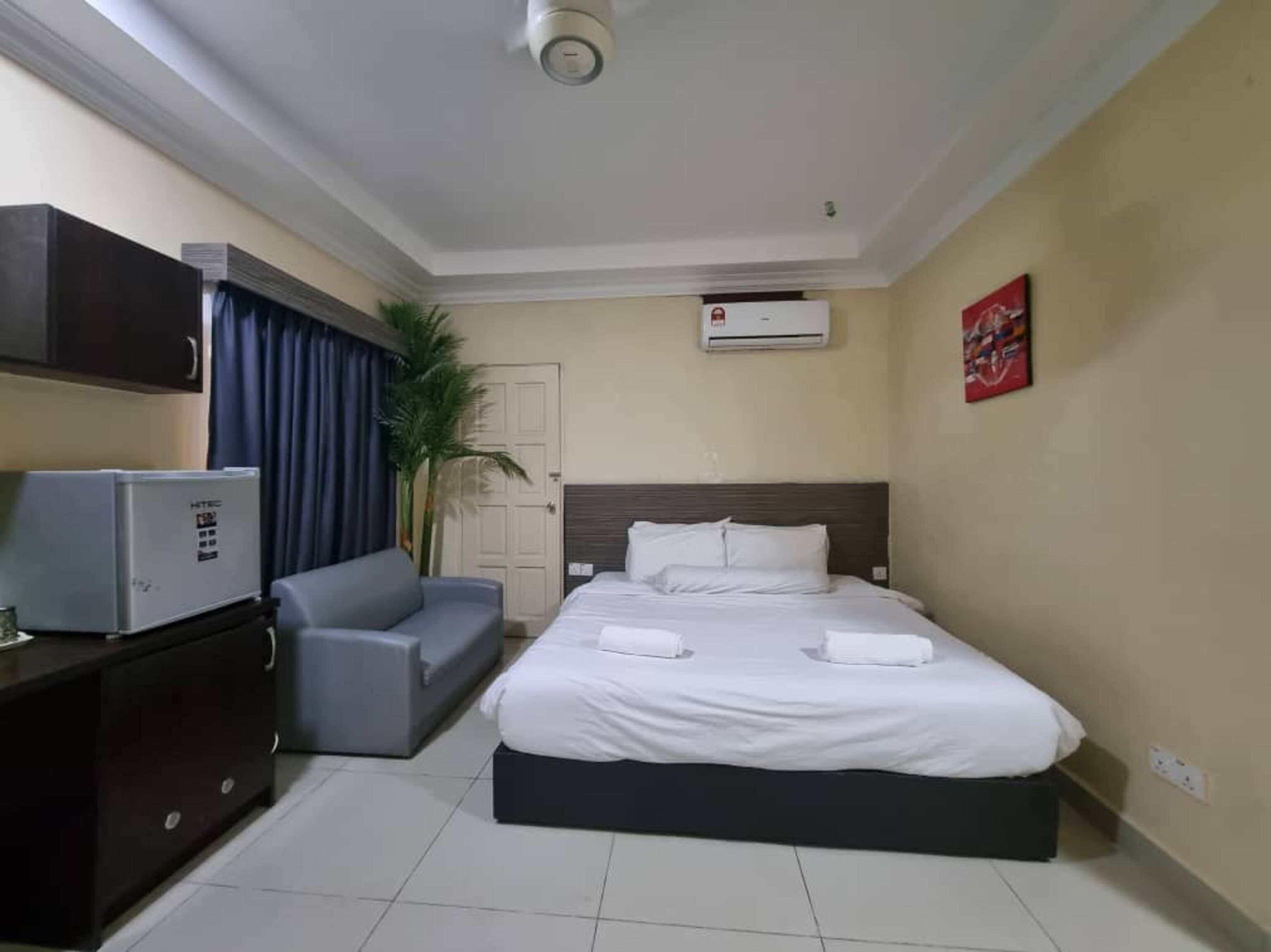 A hotel room with a bed, a desk with a refrigerator, a chair, a potted plant, and an air conditioning unit.