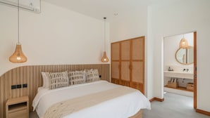 A bedroom with a wooden headboard, a bed with white bedding, two hanging light fixtures, a wooden nightstand, a wardrobe with a woven door, and a bathroom with a mirror and shelves. - Villa Honey (Canggu)
