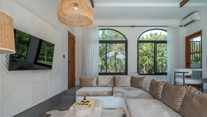 A modern living room with a sectional sofa, a flat-screen TV mounted on a textured wall, and large windows offering a view of greenery. - Villa Honey (Canggu)