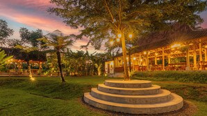 A well-lit outdoor area with a circular stone structure, surrounded by lush greenery and a building with a tiled roof.