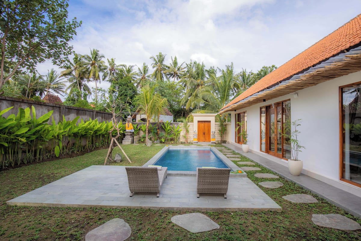 A swimming pool with two wicker chairs, surrounded by a garden with palm trees and a stone pathway.