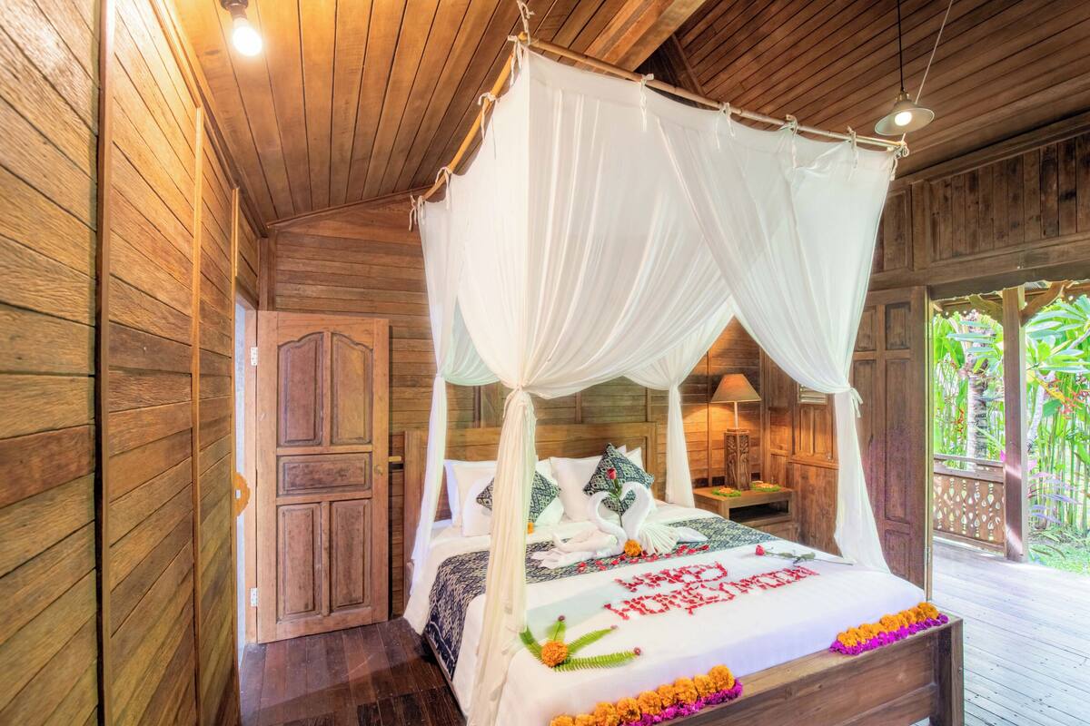 A wooden room with a four-poster bed, a wooden wardrobe, and a balcony with a view of greenery.