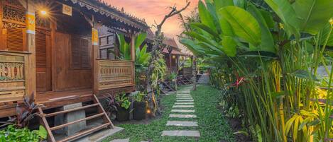 A wooden house with a thatched roof, surrounded by lush greenery and a stone pathway.