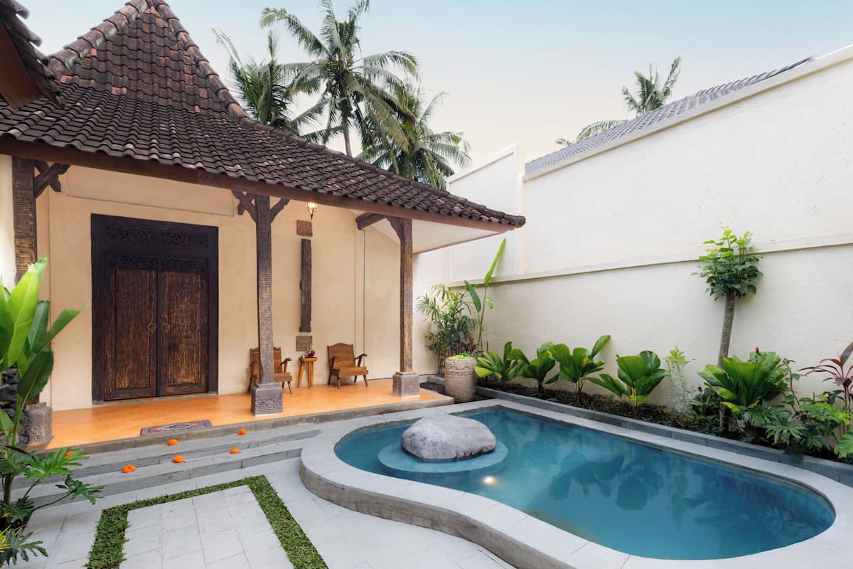 A swimming pool with a stone centerpiece, surrounded by a garden with various plants and a traditional tiled roof structure.