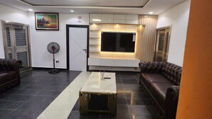 A modern living room with a black and white tiled floor, a brown leather sofa, a white coffee table, a standing fan, and a TV mounted on the wall. - Cozy Residence (Abuja)