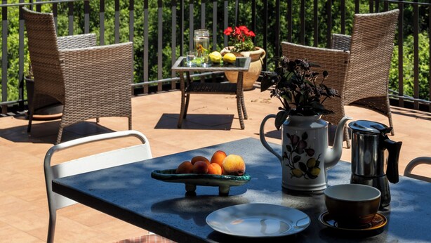 A terrace with wicker chairs, a table with fruits, a vase with flowers, a teapot, and a view of a hill with a building.
