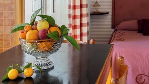 A room with a bed, a table with a fruit bowl, a window with curtains, and a wall with a sun decoration.