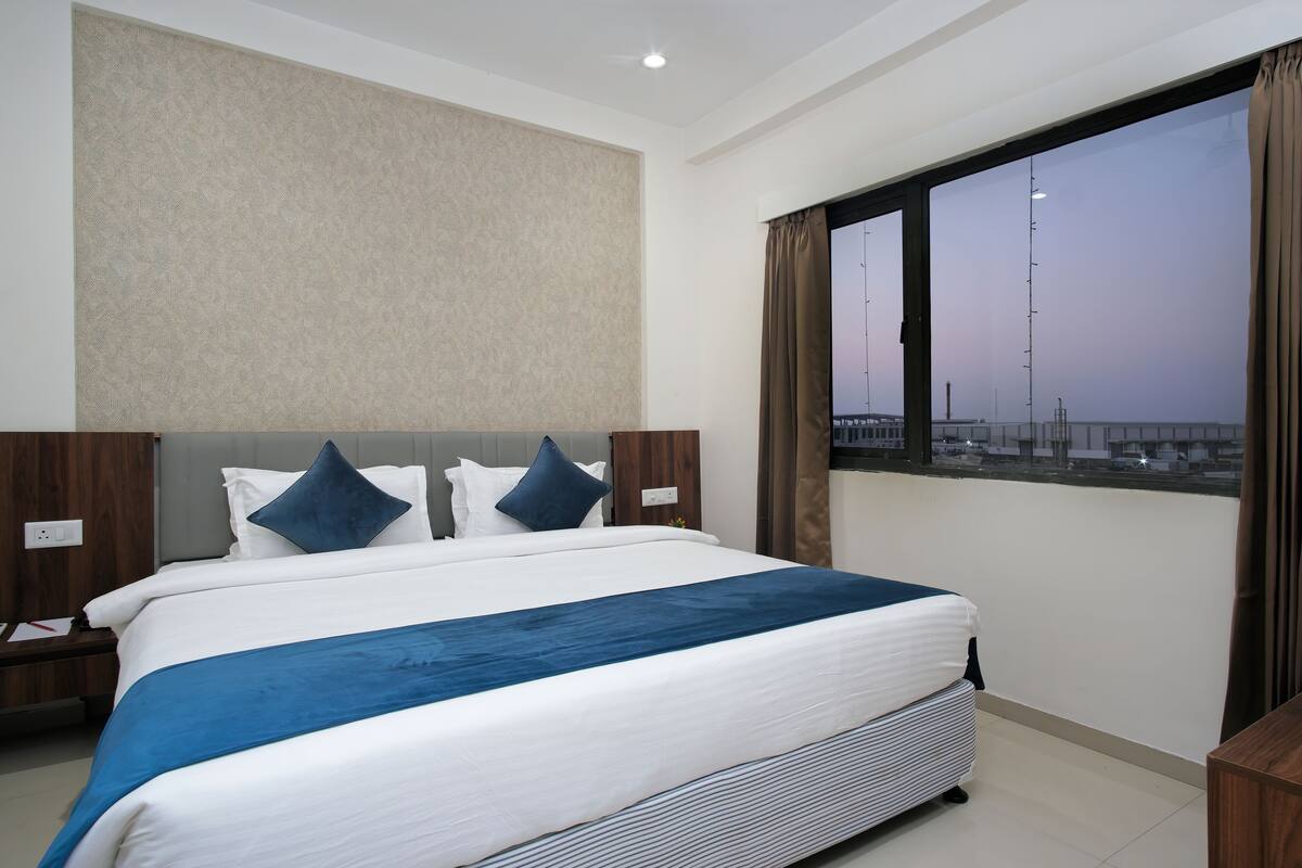 Una cama bien hecha con un detalle azul, un ventanal con vistas a una construcción distante y una pared lisa con textura.