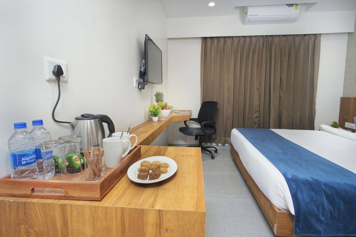 Una habitación de hotel con cama, escritorio con cafetera, silla, televisor montado en la pared y ventana con cortinas.