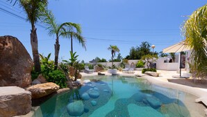 Una piscina cristalina con rocas, palmeras y sillas de descanso.