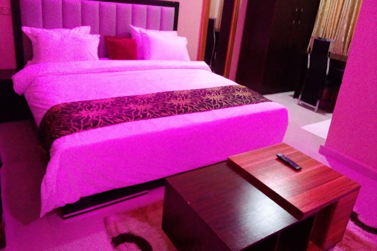 A bed with a headboard, pillows, and a blanket. There is a wooden nightstand with a remote control on it.