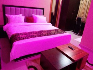 A bed with a headboard, pillows, and a blanket. There is a wooden nightstand with a remote control on it. - Prismar International Hotel (Uyo)