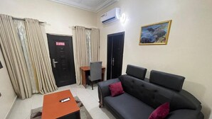 A living room with a sofa, a coffee table, a desk, and a chair. There is a door with a sign, an air conditioning unit, and a painting on the wall. - Prismar International Hotel (Uyo)