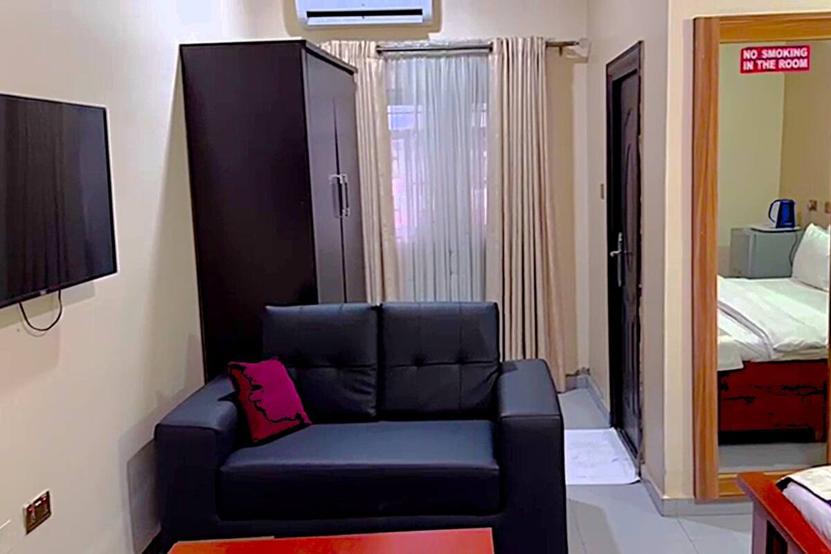A room with a black sofa, a TV mounted on the wall, a wardrobe, and a bed with a red headboard.