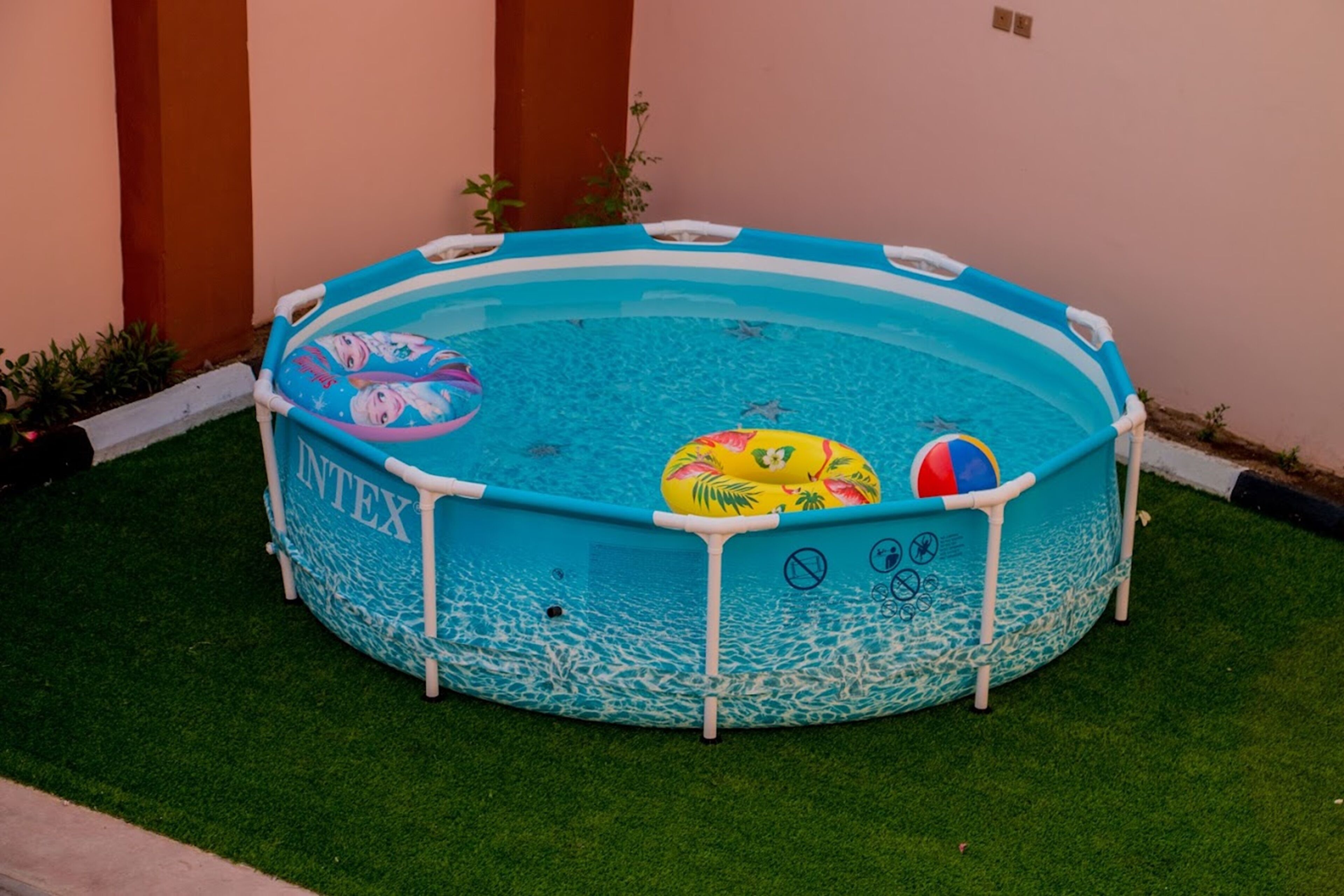 Una piscina inflable pequeña con diseño azul y blanco, rodeada de césped artificial y una pared de fondo.