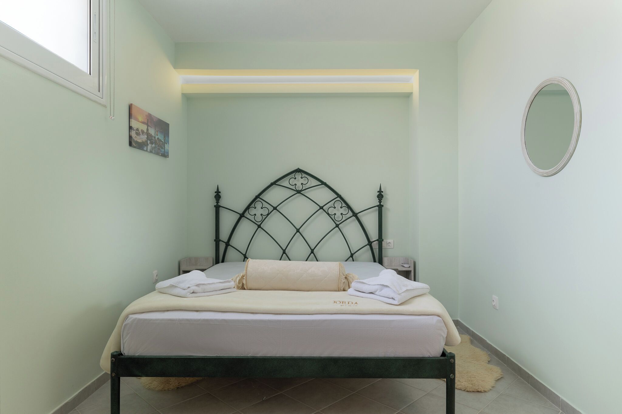 A neatly made bed with a metal headboard, a bedside table, a wall-mounted mirror, and a small picture on the wall.