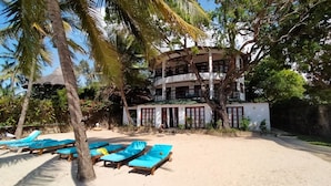 A beachfront hotel with multiple floors, a sandy beach with blue lounge chairs, and palm trees. - Silver sand beach hotel (Malindi)