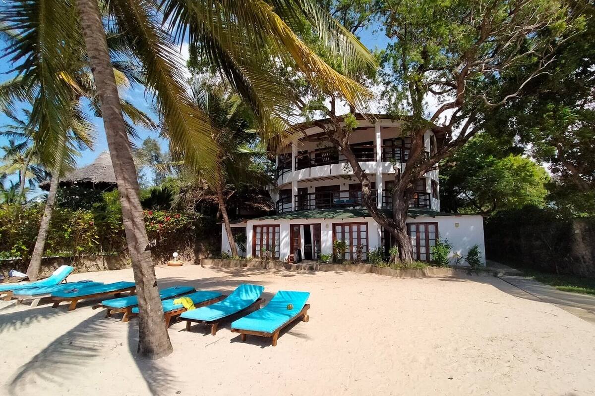 A beachfront hotel with multiple floors, a sandy beach with blue lounge chairs, and palm trees.