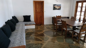 A room with a stone floor, a wooden bench with black cushions, a wooden door, a dining table with chairs, and a painting on the wall. - Silver sand beach hotel (Malindi)