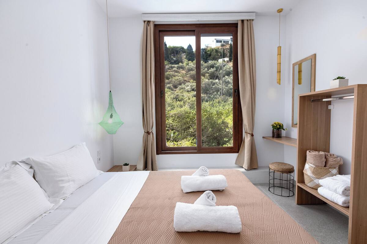 Ein Hotelzimmer mit Bett, einem Vorhangfenster, einer Pendelleuchte, einem Holzregal mit Handtüchern und einem kleinen Hocker.