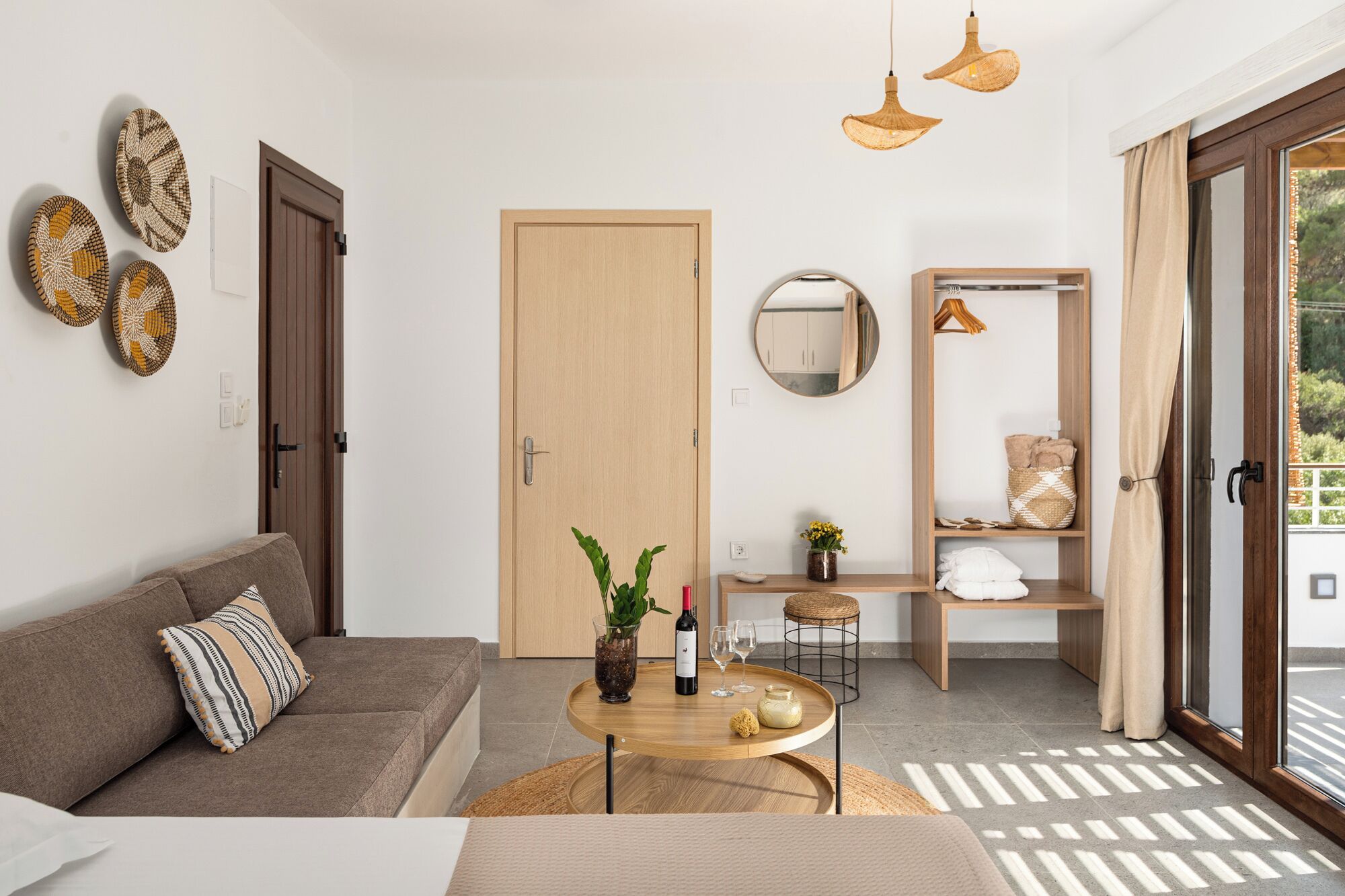 A modern living room with a sofa, a coffee table, and a dining area. There is a door, a mirror, and a shelf with towels and a basket.
