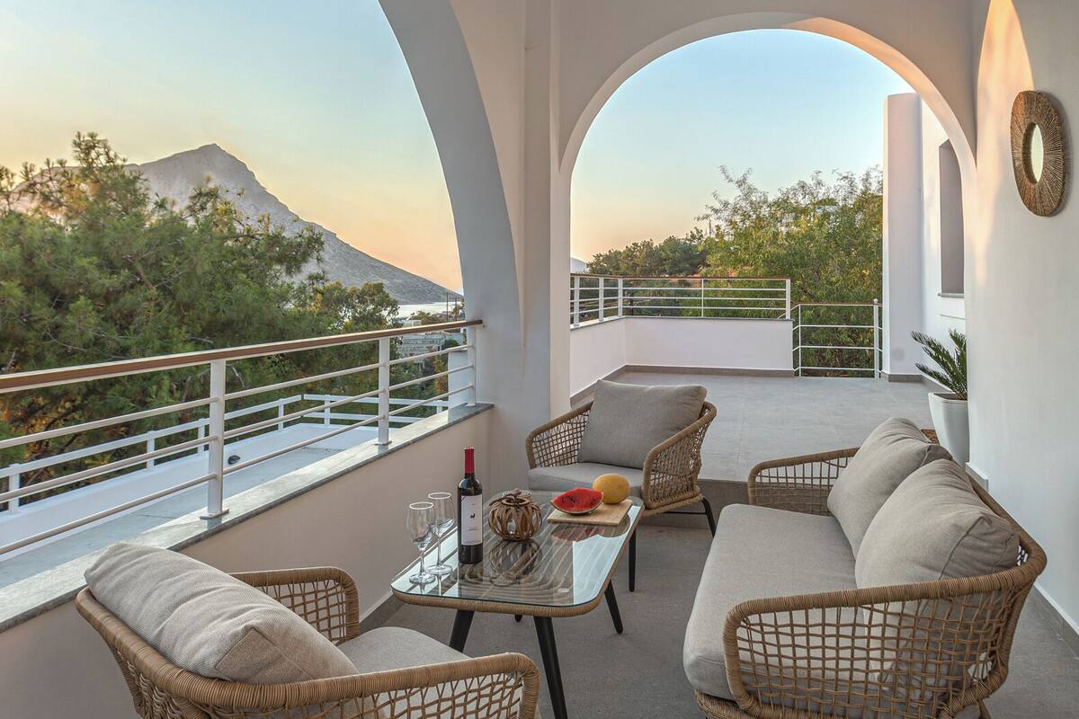 Ein Balkon mit Korbmöbeln, einem Tisch mit Flasche und Gläsern sowie Blick auf einen Berg und Bäume.