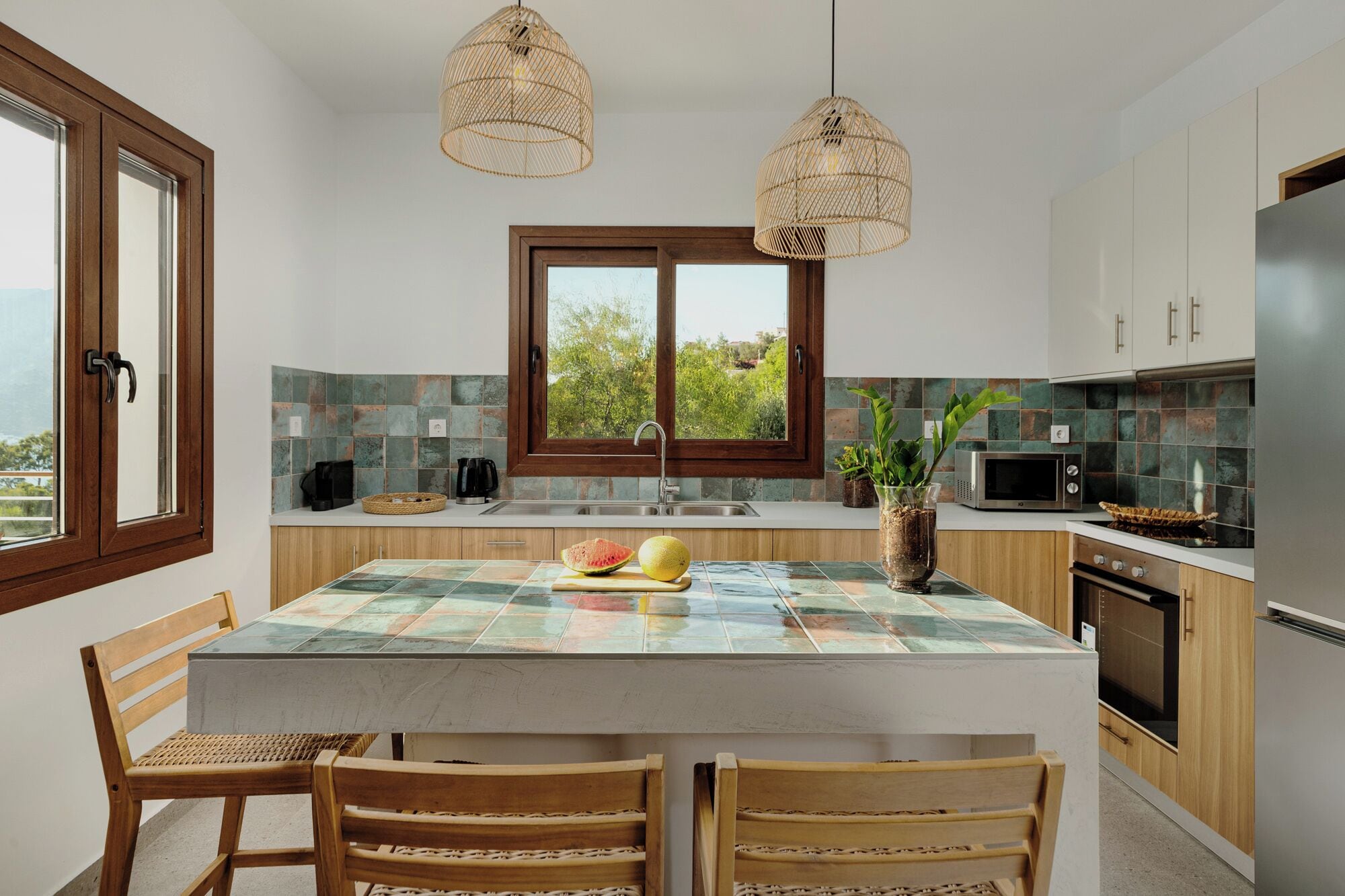 A modern kitchen with a central island, wooden chairs, a large window, and a view of greenery outside.