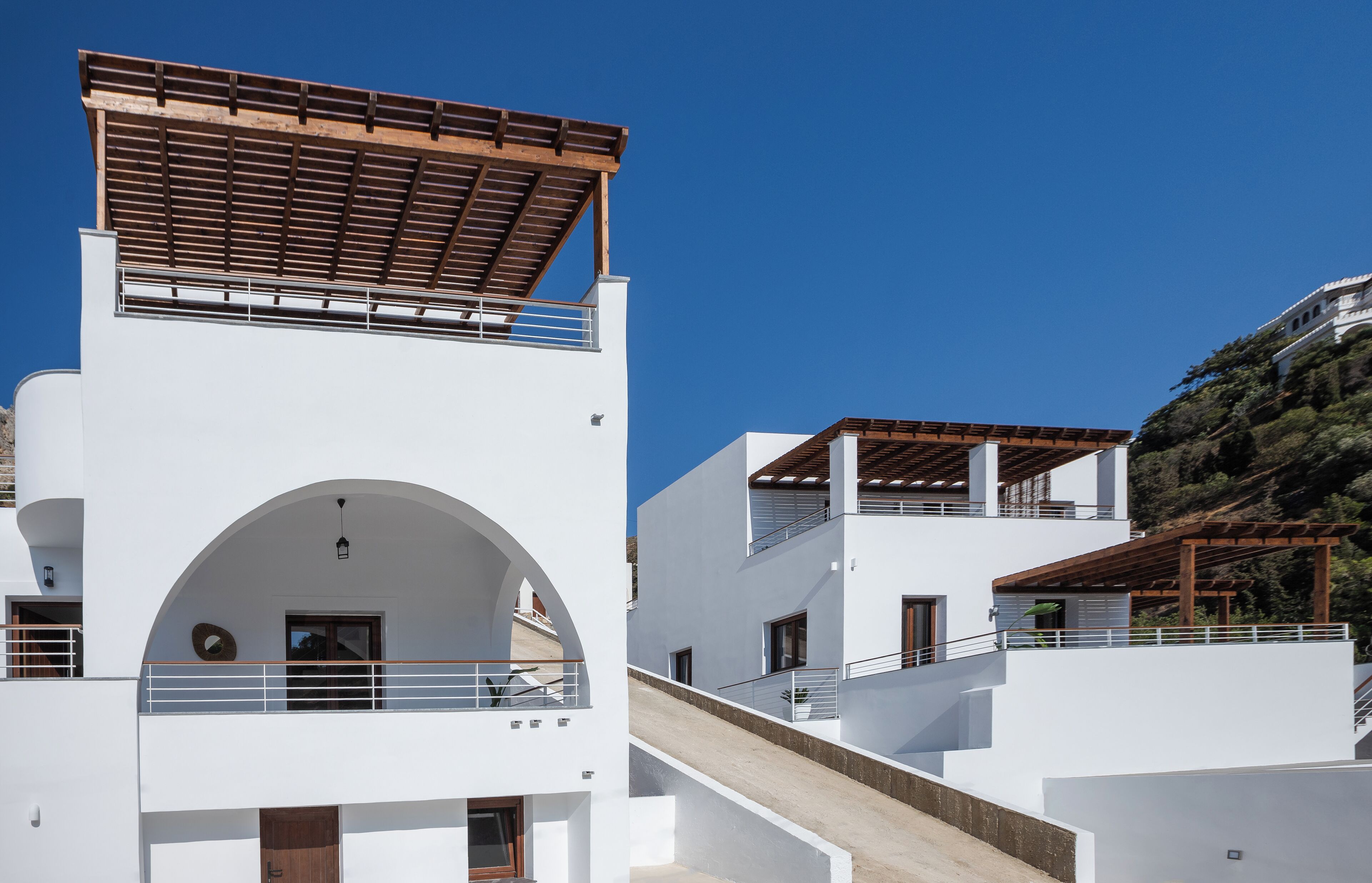 Un edificio moderno blanco con una pérgola de madera en el piso superior, una escalera que conduce al nivel superior y vistas a una colina.