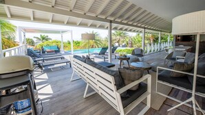 A covered outdoor seating area with multiple lounge chairs and sofas, a pool in the background, and a white ceiling structure. - Spacious Villa Near Beach (Jan Thiel)