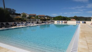 Una piscina rectangular con agua azul cristalina, rodeada de sillas de descanso y sombrillas, y un área de terraza con baldosas.