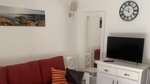 Una sala moderna con un sofá rojo, una mesa de centro blanca, un televisor sobre un mueble blanco, un reloj de pared y un cuadro de un paisaje.