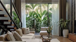 A modern living room with a sofa set, a coffee table, and a large window with a view of tropical plants. - Sunny Cuddles (Canggu)
