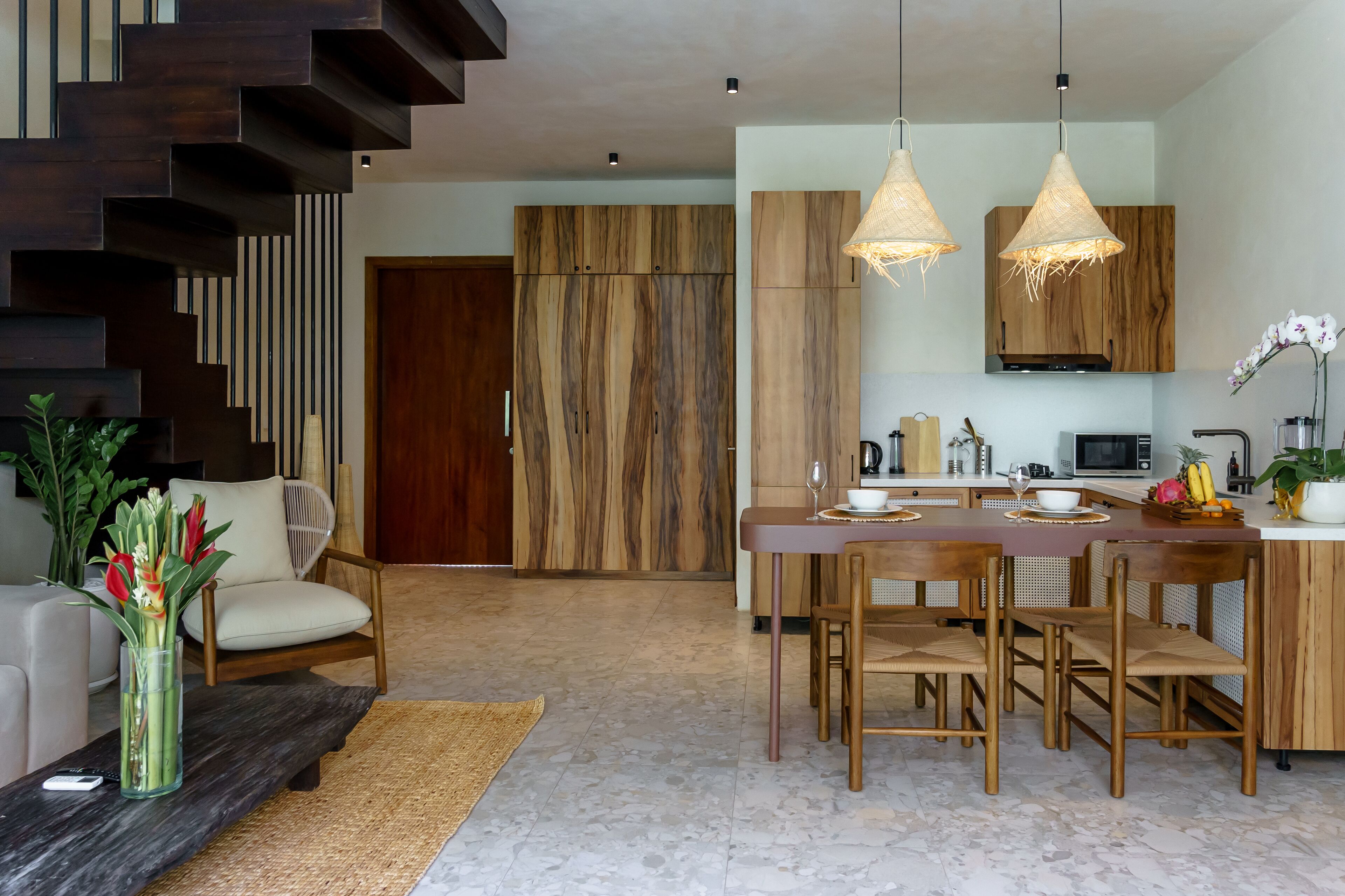 A modern kitchen with wooden cabinets, a dining area with a wooden table and chairs, and a staircase with dark wood finishes.