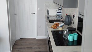 Eine kleine Küche mit weißer Tür, einem Spülbecken, einem Herd und einer Arbeitsplatte mit Wasserkocher, Croissants und einer Kaffeemaschine.