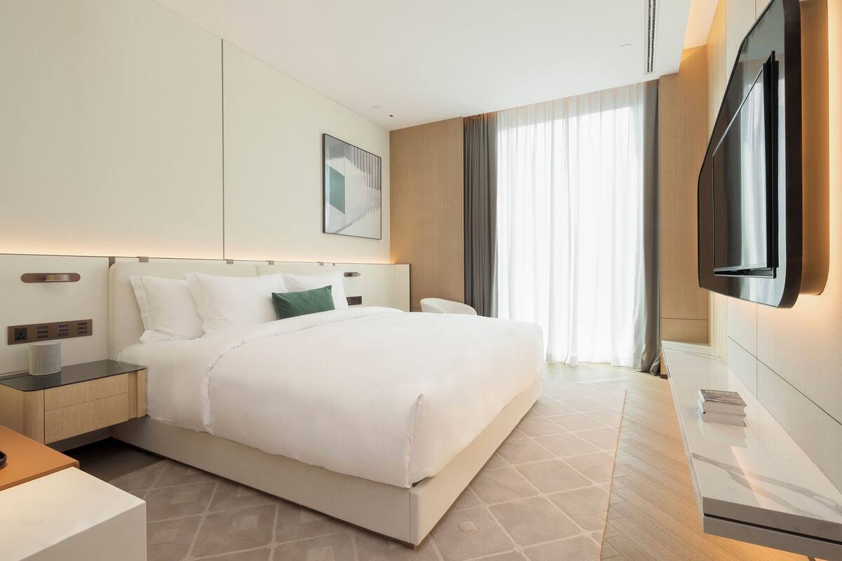 A modern hotel room with a large bed, bedside tables, a flat-screen TV, and a large window with curtains.