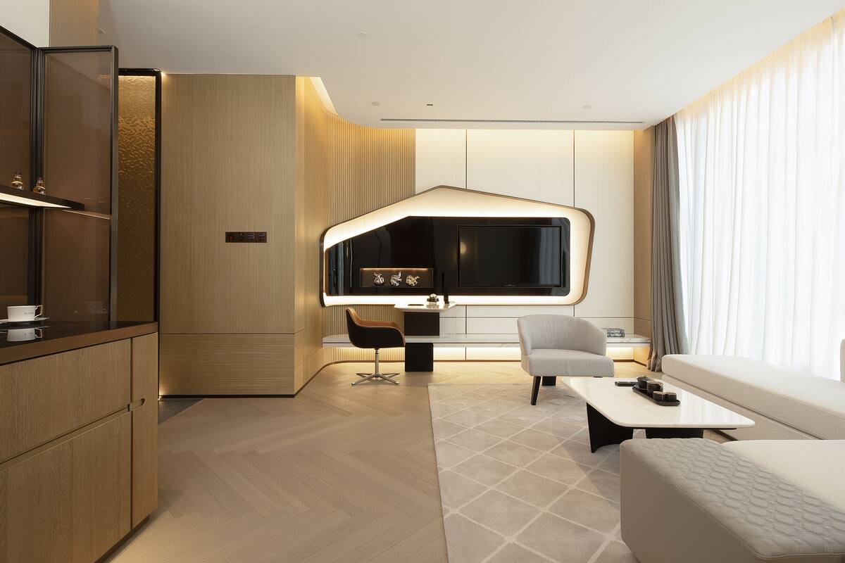 A modern hotel room with a kitchen area, a dining table, a sofa, and a large window with curtains.