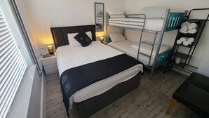 Een kleine hotelkamer met een stapelbed, een eenpersoonsbed, een nachtkastje met lamp, een spiegel en een plank handdoeken.