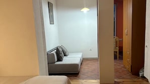 A bedroom with a wooden floor, a bed, a sofa, and a small dining area visible through an open doorway. - Themis Apart Hotel (Kotor)