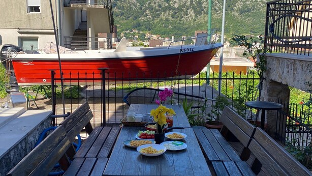 A wooden deck with a table set for a meal, overlooking a red boat and a mountainous landscape. - Themis Apart Hotel (Kotor)