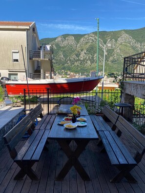 A wooden deck with a table set for a meal, overlooking a red boat and a mountainous landscape. - Themis Apart Hotel (Kotor)