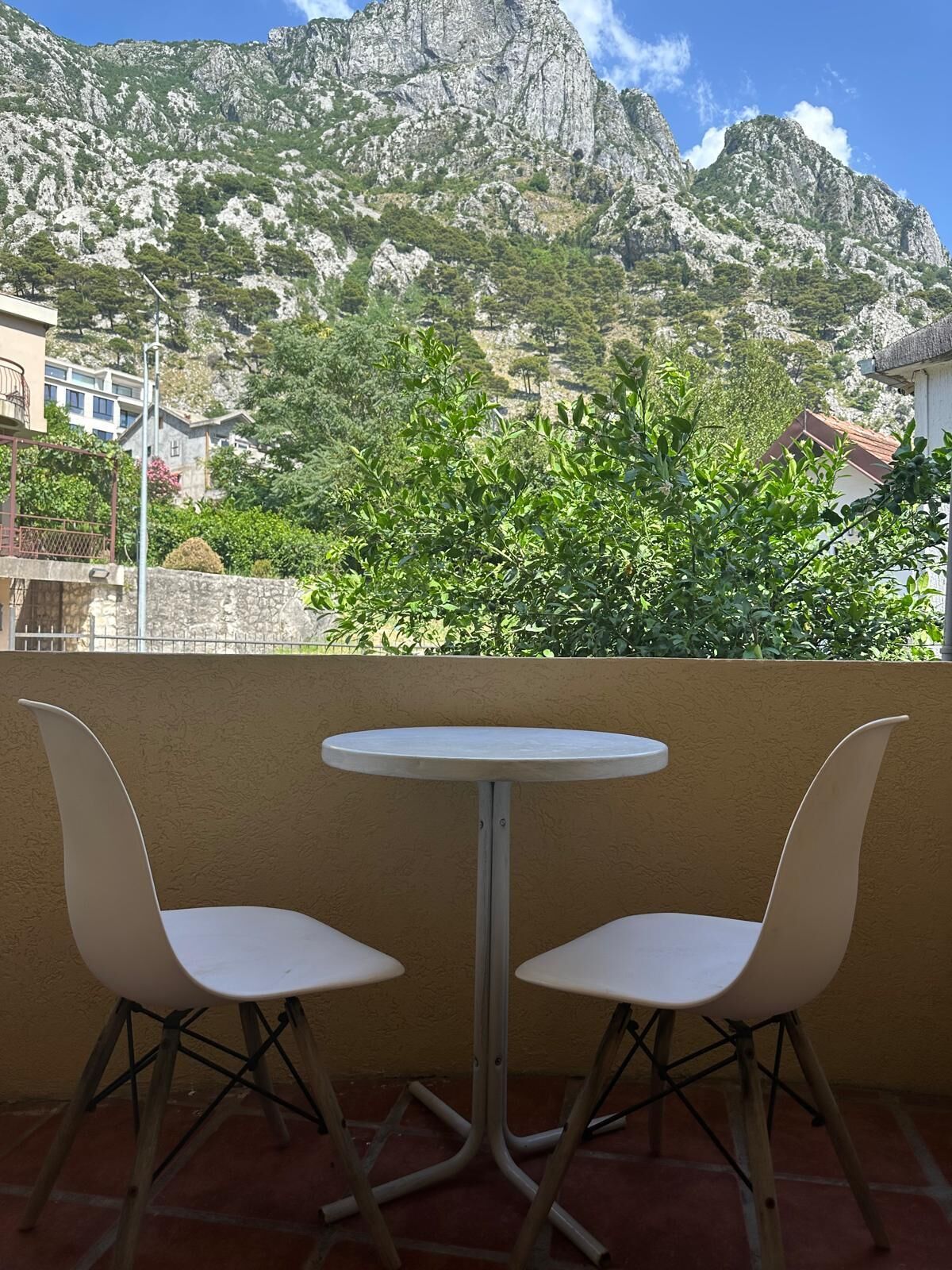 A balcony with two white chairs and a small round table, overlooking a mountainous landscape with greenery and buildings in the background.
