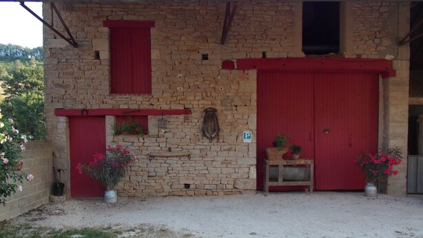 A stone building with a red door and window shutters, a small table with potted plants, and a gravel driveway. - Gite a la Campagne (La Roche-Vanneau)