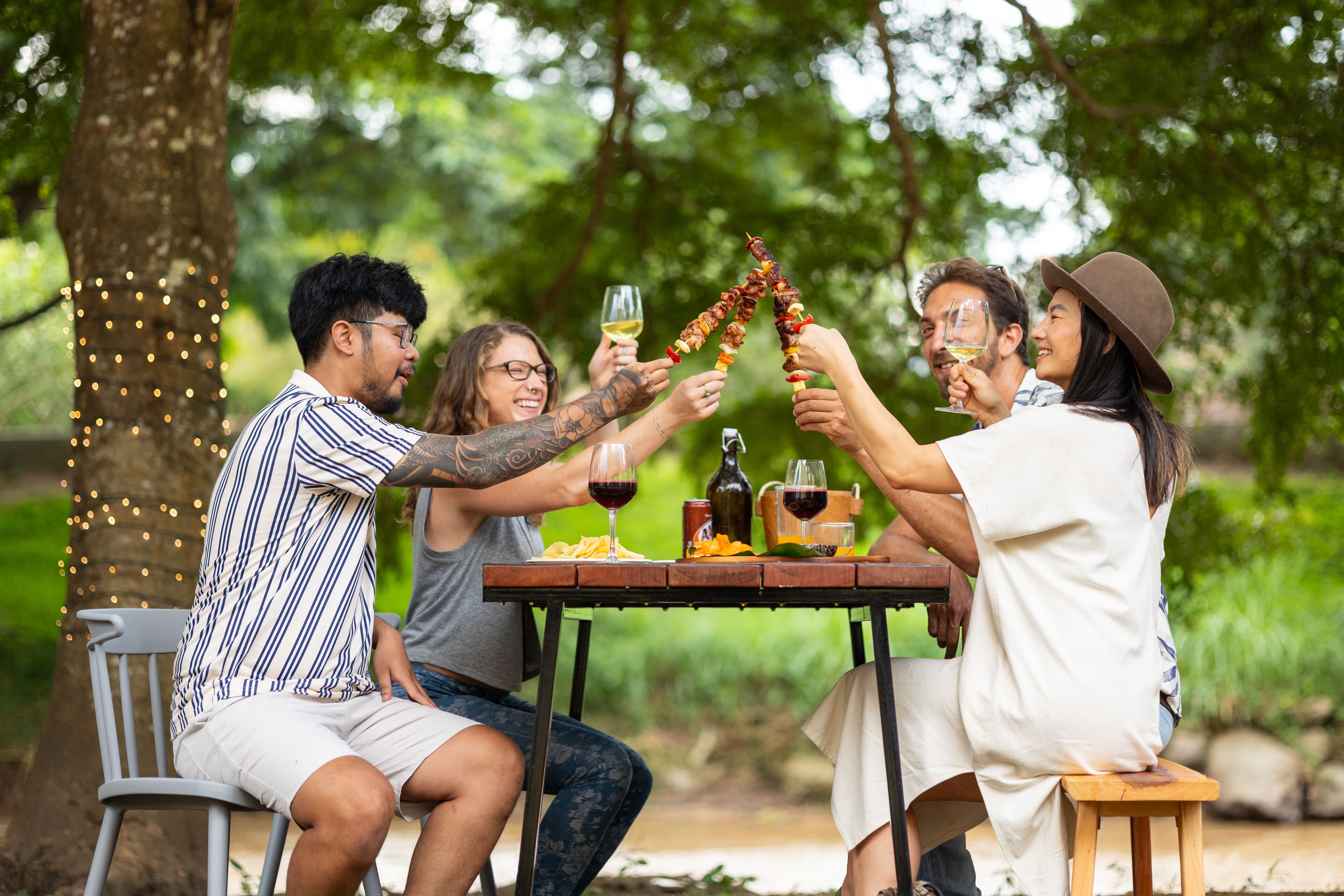 A group of people toasting with skewers and wine glasses at an outdoor table with a view of greenery.
